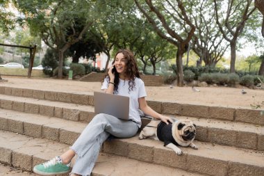 Keyifli esmer serbest çalışan, gündelik giysiler içinde laptopun yanında akıllı telefondan konuşan ve İspanya 'nın Barcelona kentindeki bulanık parkta vakit geçirirken merdivenlerde köpek besleyen biri. 