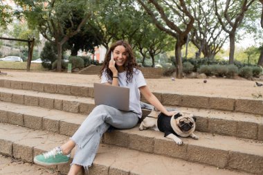 Positive and curly young freelancer talking on smartphone near laptop, looking at camera and petting pug dog while sitting on stairs in park in Barcelona, Spain  clipart