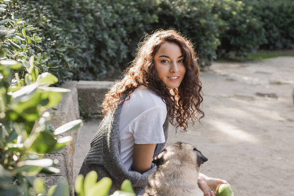 Carefree young curly woman in t-shirt and sweater holding green apple and looking at camera while sitting on stone bench near pug dog and relaxing in park in Barcelona, Spain 