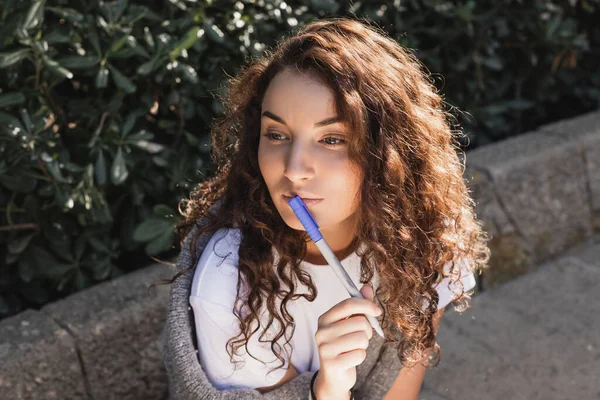 stock image Pensive and young curly woman in casual clothes holding marker near lips and looking away while sitting on blurred stone bench in park at daytime in Barcelona, Spain, sunny day