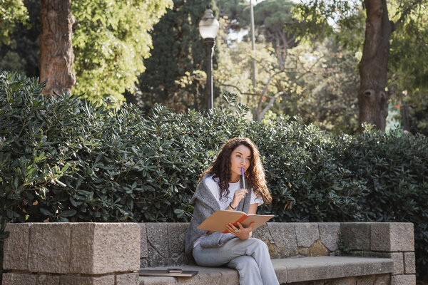 Pensive young and curly woman in casual clothes holding notebook and marker near lips while sitting near devices on stone bench and green plants in park at daytime in Barcelona, Spain 