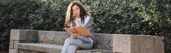 Focused young curly woman looking at camera while holding marker and notebook near smartphone and laptop on stone bench near bushes in park in Barcelona, Spain, banner, work from anywhere 