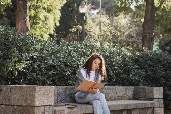 Young curly woman in casual clothes holding notebook while sitting near devices on stone bench and green bushes and spending time in park at daytime in Barcelona, Spain 