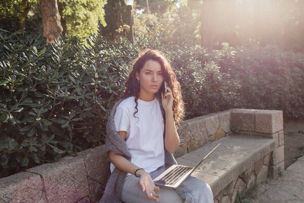 Pensive young and curly freelancer in casual clothes talking on smartphone near laptop while sitting on stone bench and spending time near green plants in park in Barcelona, Spain 