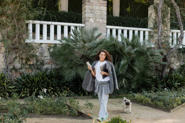 stock image Cheerful and curly freelancer in warm jacket and earphones looking at camera and holding devices while walking near pug dog in park at daytime in Barcelona, Spain 