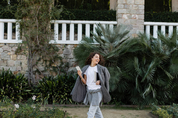 Carefree brunette freelancer in warm jacket listening music in earphones and holding laptop with smartphone while walking in park at daytime in Barcelona, Spain 