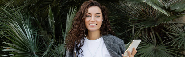 Positive and young curly freelancer in earphones and casual clothes holding gadgets and looking at camera while standing near palm trees in park at daytime in Barcelona, Spain, banner 