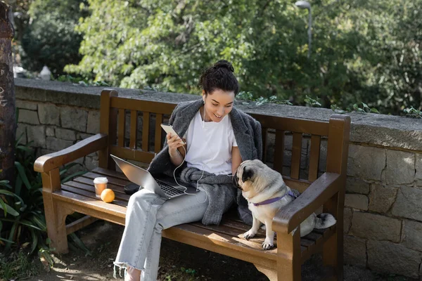 stock image Smiling freelancer in earphones holding smartphone and looking at pug dog while sitting near laptop, coffee to go and orange on wooden bench and spending time in park in Barcelona, Spain 