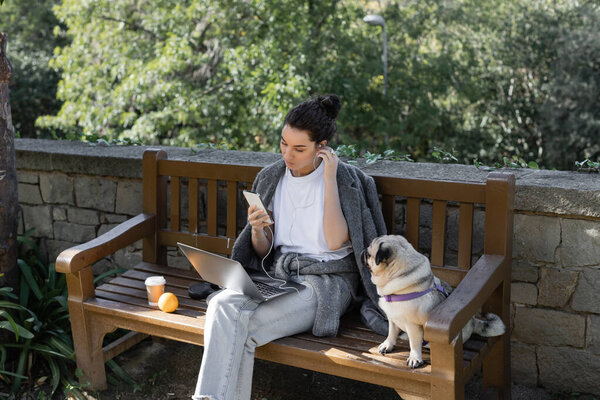 Young and curly freelancer listening music in earphones, using smartphone near laptop and sitting next to pug dog, orange and coffee to go on wooden bench in park in Barcelona, Spain 