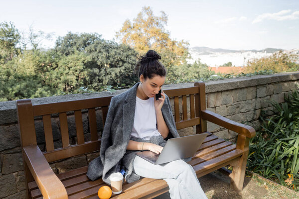 Young brunette freelancer in warm jacket talking on smartphone and using laptop while sitting near coffee to go and orange on wooden bench in park in Barcelona, Spain, work-life balance 