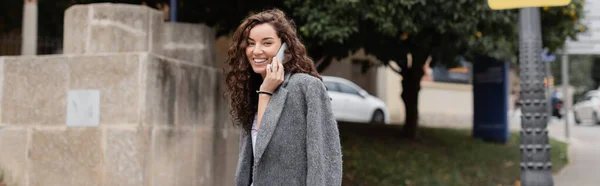 ポジティブな若いと巻きブルネットの女性でカジュアルなジャケットカメラを見て携帯電話で話す一方 バルセロナ スペイン バナーのぼやけた都市部の通りに立って — ストック写真