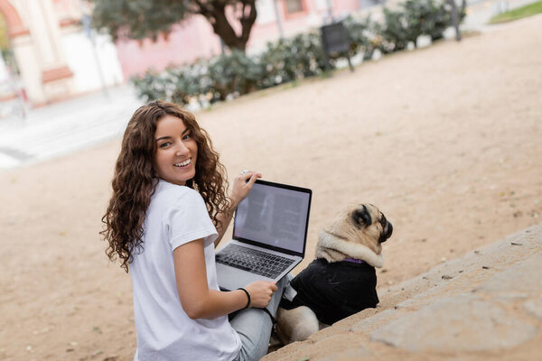Cheerful young curly woman in casual clothes looking at camera while holding laptop near pug dog on stairs in blurred park in Barcelona, Spain, white t-shirt 