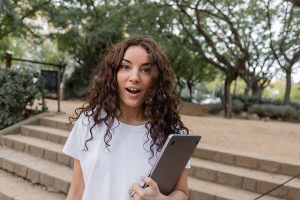 Shocked young and curly freelancer in white t-shirt looking at camera with open mouth while holding laptop and spending time in blurred park at daytime in Barcelona, Spain, 