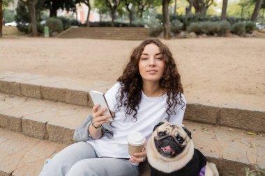 Barcelona, İspanya 'da bulanık bir parkta merdivenlerde oturan Pug Dog' un yanındaki kameraya bakarken günlük giysiler içinde gülümseyen genç kıvırcık kadın cep telefonu kullanıyor ve kahve tutuyor. 