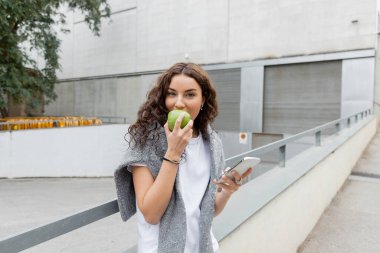 Kıvırcık ve esmer kadın kameraya bakarken taze yeşil elma yiyor ve İspanya 'nın Barselona kentsel caddesinde bulanık sanayi binasının yanında akıllı telefon kullanıyor. 