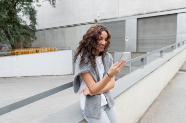 Gülümseyen esmer kadın omuzlarında kalın bir kazakla akıllı telefon kullanarak Barcelona, İspanya 'da gündüz vakti bulanık şehir sokağında yeşil ağaçla dikiliyor.