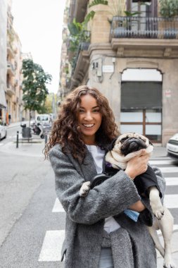 Sıcak ceketli neşeli genç kadın kameraya bakıyor ve pug dog 'u elinde tutuyor. İspanya' nın Barselona kentindeki şehir caddesinde gündüz vakti bulanık bir binanın yanında duruyor. 