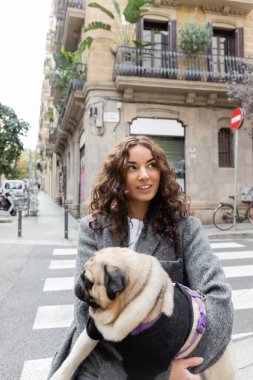 Neşeli ve kıvırcık montlu genç kadın pug dog tutuyor ve bulanık şehir sokağında gündüz vakti İspanya 'nın Barselona şehrinde binalarla dikilirken başka yere bakıyor. 