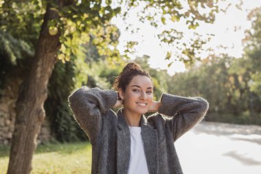 Pretty brunette woman in casual clothes and jacket touching hair and smiling at camera while spending time in blurred green park at daytime in Barcelona, Spain  clipart