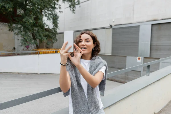 ケアフリー若いですブルネット女性とともに暖かい灰色のセーター肩の上にビデオ通話を持っていますスマートフォン上で立ちながら バルセロナ スペインの背景に工業ビルと都市の通りに — ストック写真