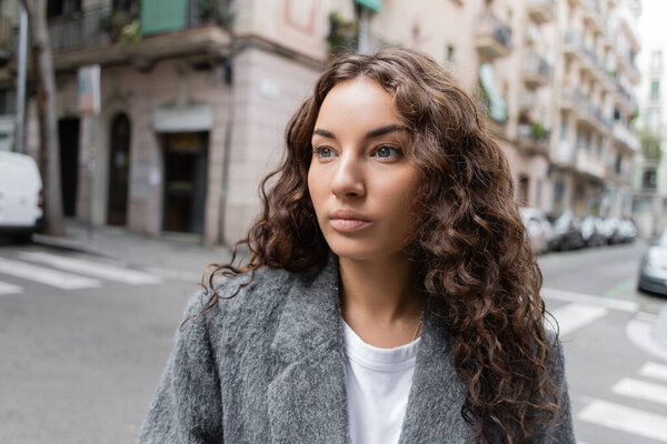 Brunette and pretty young woman in casual jacket looking away while standing on blurred city street with buildings at background in Barcelona, Spain 