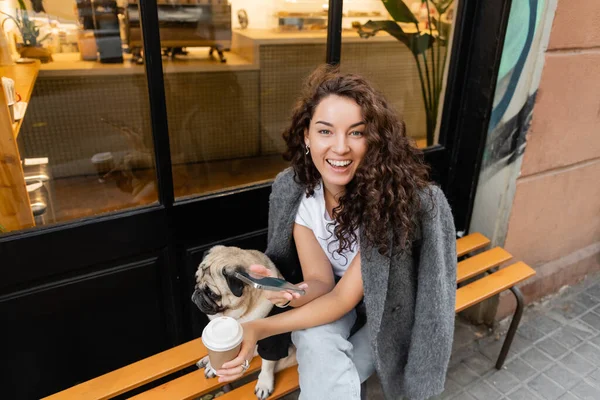 Barcelona, İspanya 'da şehir sokağındaki açık kafenin yanındaki bankta otururken elinde akıllı telefon ve kahve tutan rahat ceketli genç esmer kadın. 