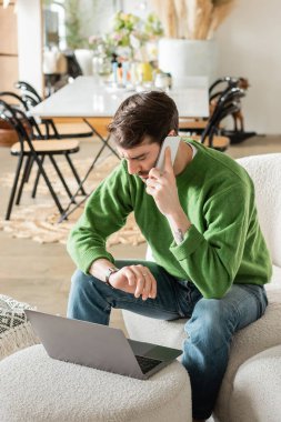 Yeşil kazaklı ve kot pantolonlu bir adam kol saatine bakıyor ve dizüstü bilgisayarla çalışırken akıllı telefondan konuşuyor. 