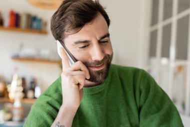 Gülümseyen sakallı ve dövmeli yeşil kazaklı adam akıllı telefondan konuşuyor ve eve bakıyor. 