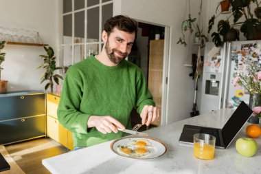 Yeşil kazaklı neşeli sakallı adam elinde çatal bıçak takımıyla modern mutfakta kahvaltı hazırlıyor. 