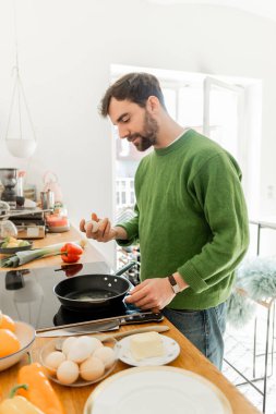 Bearded man in jumper holding egg near frying pan, butter and fresh food on worktop  clipart