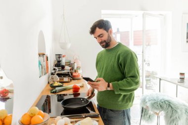 Sakallı adam akıllı telefon ve tavayı yumurta, taze yemek ve tereyağı yanında tutuyor.