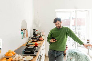 Sakallı adam akıllı telefon kullanıyor ve modern mutfakta atölyenin yanında duruyor. 