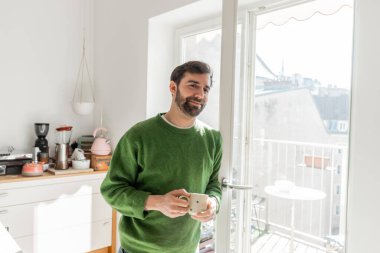 Cheerful bearded man in jumper holding cup of coffee and looking at camera  clipart