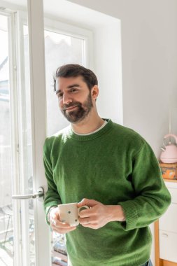 Olumlu sakallı, esmer, yeşil kazaklı bir adam elinde kahve bardağıyla pencerenin yanında duruyor.