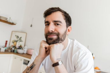 Beyaz tişörtlü, sakallı ve dövmeli bir adamın portresi kameraya bakıyor, çenesinin yanında el ele tutuşuyor. 