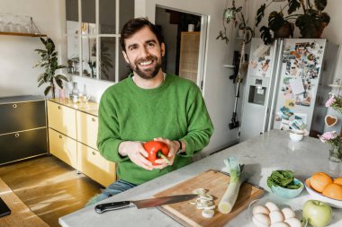 Cheerful bearded man in green jumper holding fresh bell pepper in modern kitchen at home  clipart