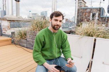 bearded man in earphones holding cup of coffee while spending time on rooftop terrace in Vienna clipart