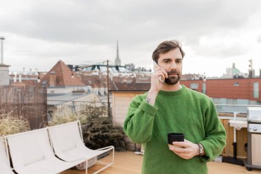 Avusturya, Viyana 'da yeşil kazaklı bir adam akıllı telefondan konuşuyor ve elinde bir fincan kahve tutuyor.
