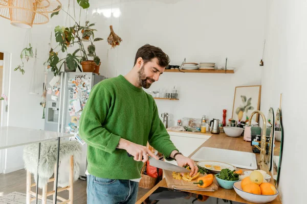 キッチンで朝食を調理しながら新鮮な唐辛子を切るジャンパーとジーンズの幸せなひげ付き男 — ストック写真