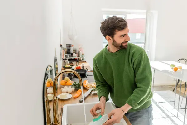 stock image Smiling bearded and tattooed man washing plate near sink and looking away at home 