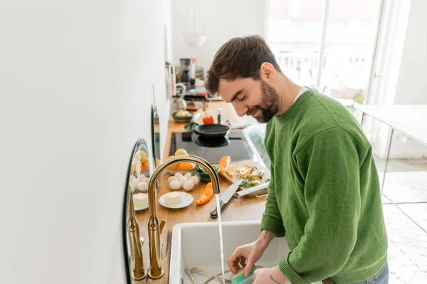 stock image Smiling and bearded man in green jumper washing tableware at home 