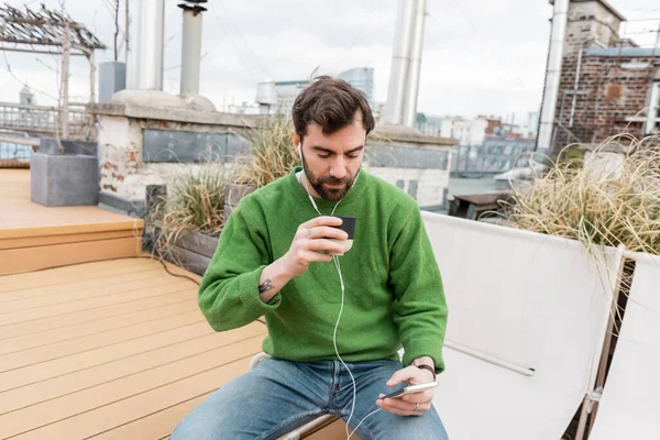 bearded man in earphones holding cup of tea while spending time on rooftop terrace in Vienna