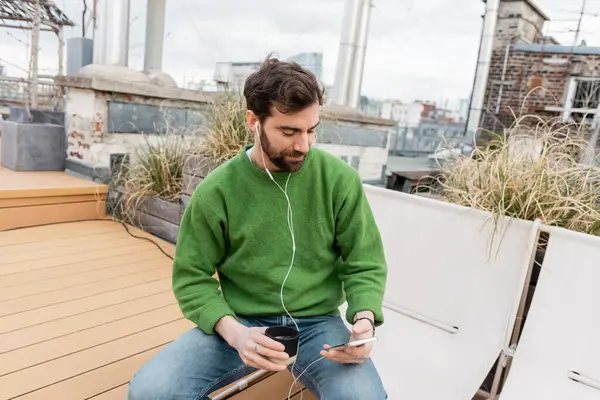 stock image bearded man listening music in earphones and holding cup of coffee on rooftop in Vienna, Austria