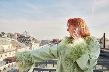 trendy woman in stylish casual attire talking on mobile phone on roof terrace in Vienna, Austria clipart