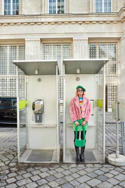 happy woman in fashionable casual attire posing in phone booth on street in Vienna, Austria