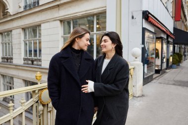 Intimate moment of cheerful lesbian women in love standing together on street in European city clipart