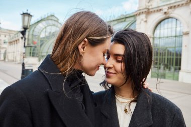 cheerful lesbian couple in coats looking at each other near Palmenhaus in Vienna on background clipart