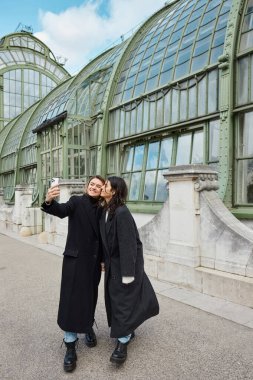 Mutlu lezbiyen çift Viyana 'da tarihi bir yerin yakınında dururken akıllı telefondan selfie çekiyor.