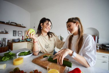 happy young lesbian couple smiling while making salad together in modern kitchen, lgbtq concept clipart