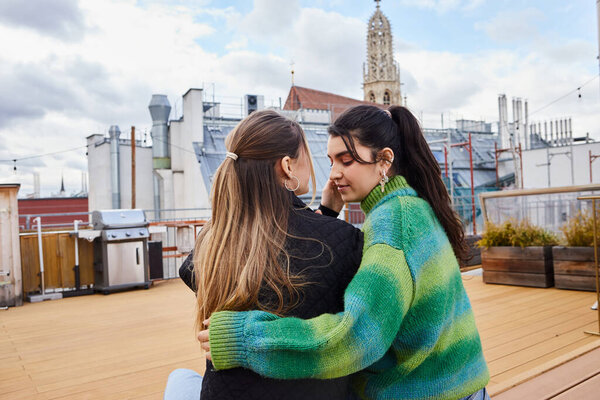 Tender moment between young lesbian women sitting together on a rooftop, cityscape backdrop